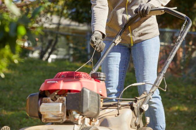 Woman wearing sweatshirt tries to start lawn mower in her yard.